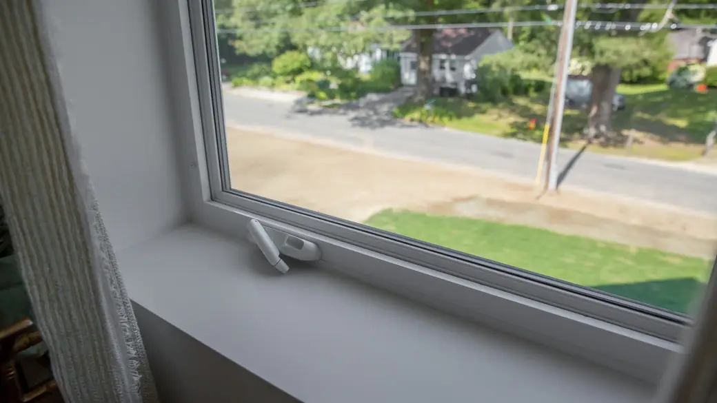 A view through a window shows a quiet suburban street, green lawns, and houses. A white plastic window lock is attached to the frame, helping to improve window energy efficiency while securing the home.