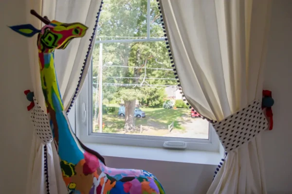 A colorful giraffe sculpture with a multicolored patchwork pattern sits on the windowsill. White curtains with black polka dot tiebacks frame my coastal windows, through which window boxes bursting with flowers add a vibrant touch against the background of a tree and lawn.