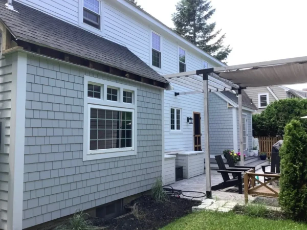 The image captures a New England backyard with light gray siding. A patio area features a wooden pergola, sheltering black chairs and a table. Verdant grass and shrubs surround the patio, reminiscent of a scene from the Farmers Almanac, while multiple windows adorn the house.