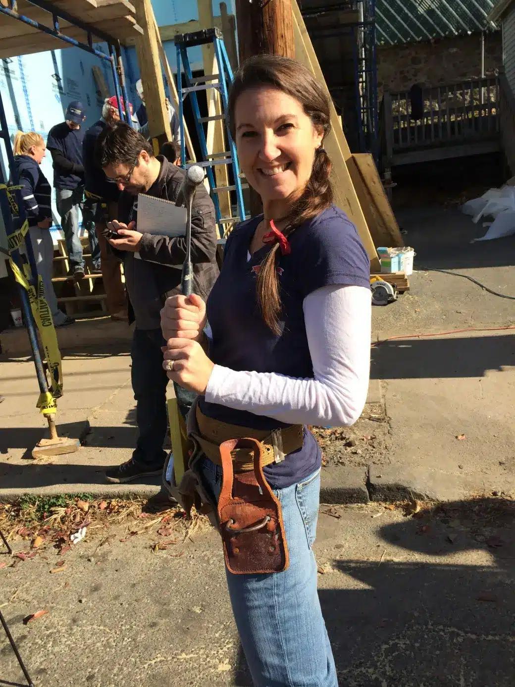 A woman with a long braid, embodying the spirit of a dedicated contractor, smiles as she holds a tool at the construction site. Dressed in a blue long-sleeve shirt and jeans with a tool belt around her waist, she stands amid fellow workers and building materials in the background.