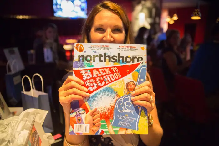 A woman holds up a North Shore Magazine Launch issue with a colorful Back to School cover, obscuring her face. The background reveals a dimly lit, busy indoor setting filled with other people and soft, ambient lights.