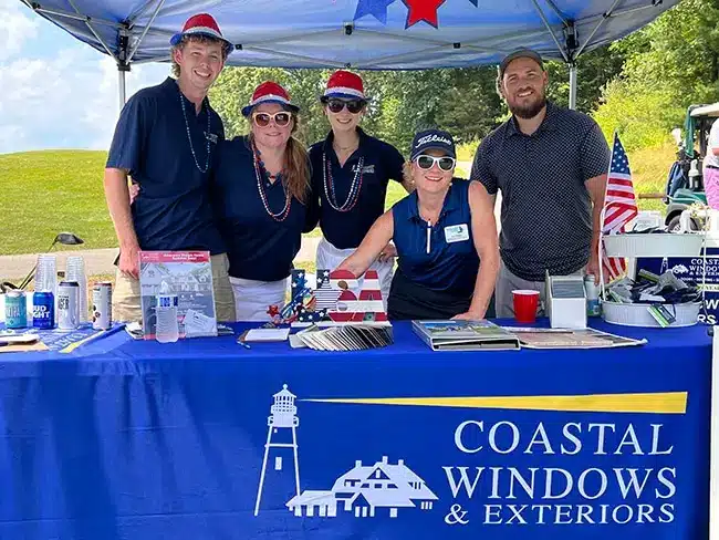 Four people stand and one sits behind a booth adorned with a Coastal Windows & Exteriors banner at the Greater Newburyport Chamber Golf Classic. Theyre dressed in patriotic-themed attire with hats, surrounded by brochures, samples, and an American flag waving gently among the trees.