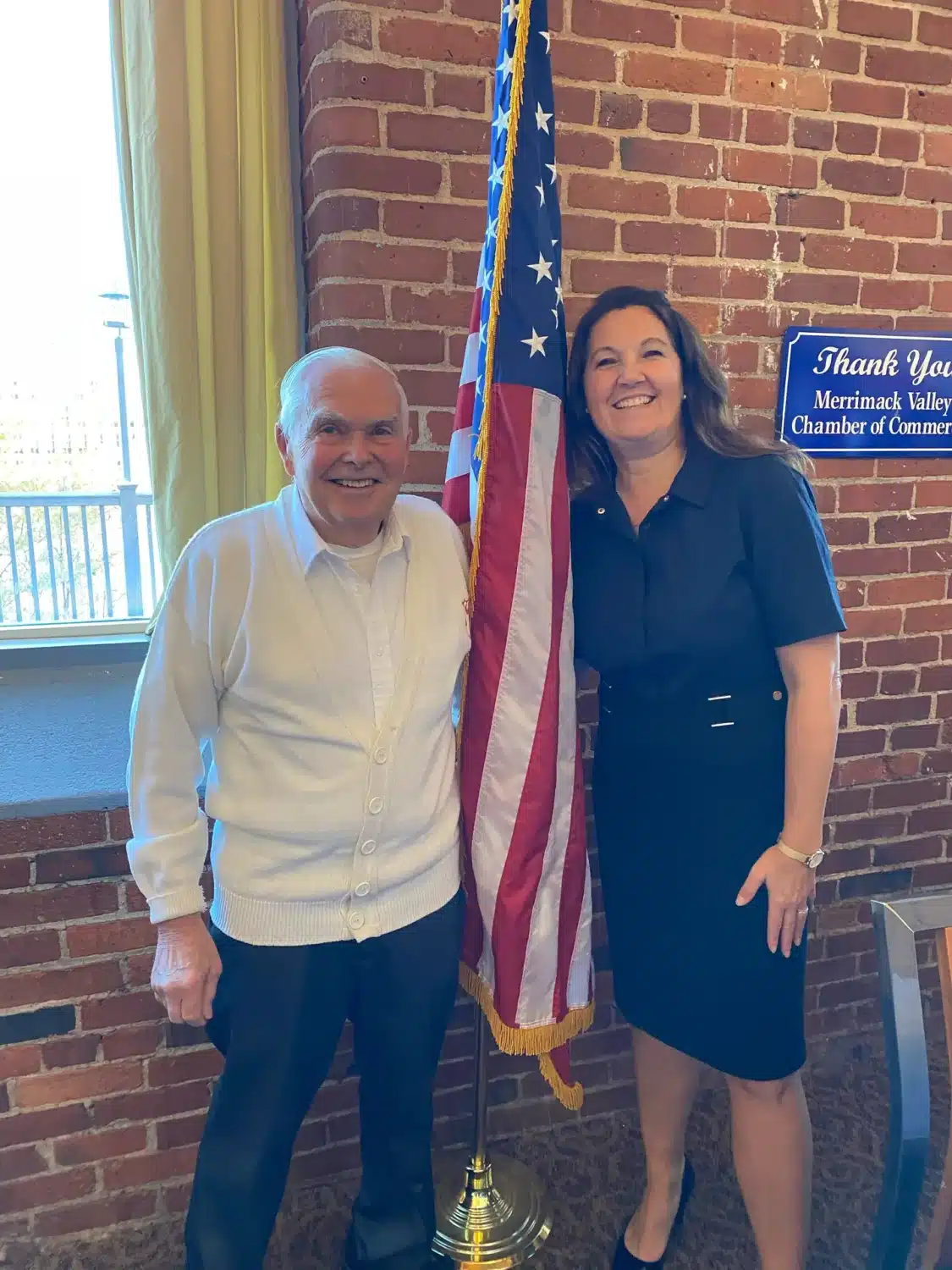 An older man in a white sweater and a woman in a dark dress stand smiling beside a U.S. flag. They are indoors, in front of a brick wall with a window and a blue sign, celebrating the Free Roof donation to the local veteran.