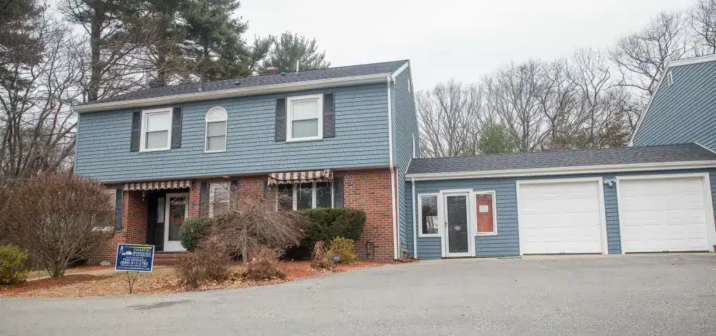 Two-story blue house with a recent home siding makeover, red brick facade, black shutters, and striped awnings over lower windows. Attached two-car garage is on the right. Leafless trees and shrubs surround the house. A for-sale sign is on the lawn.