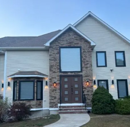 Modern two-story house with Hardie Siding in Rowley MA, a stone facade around the entrance, large square and rectangular windows, brown front door, lantern-style lights, shrubs, and a curved walkway leading to the door.