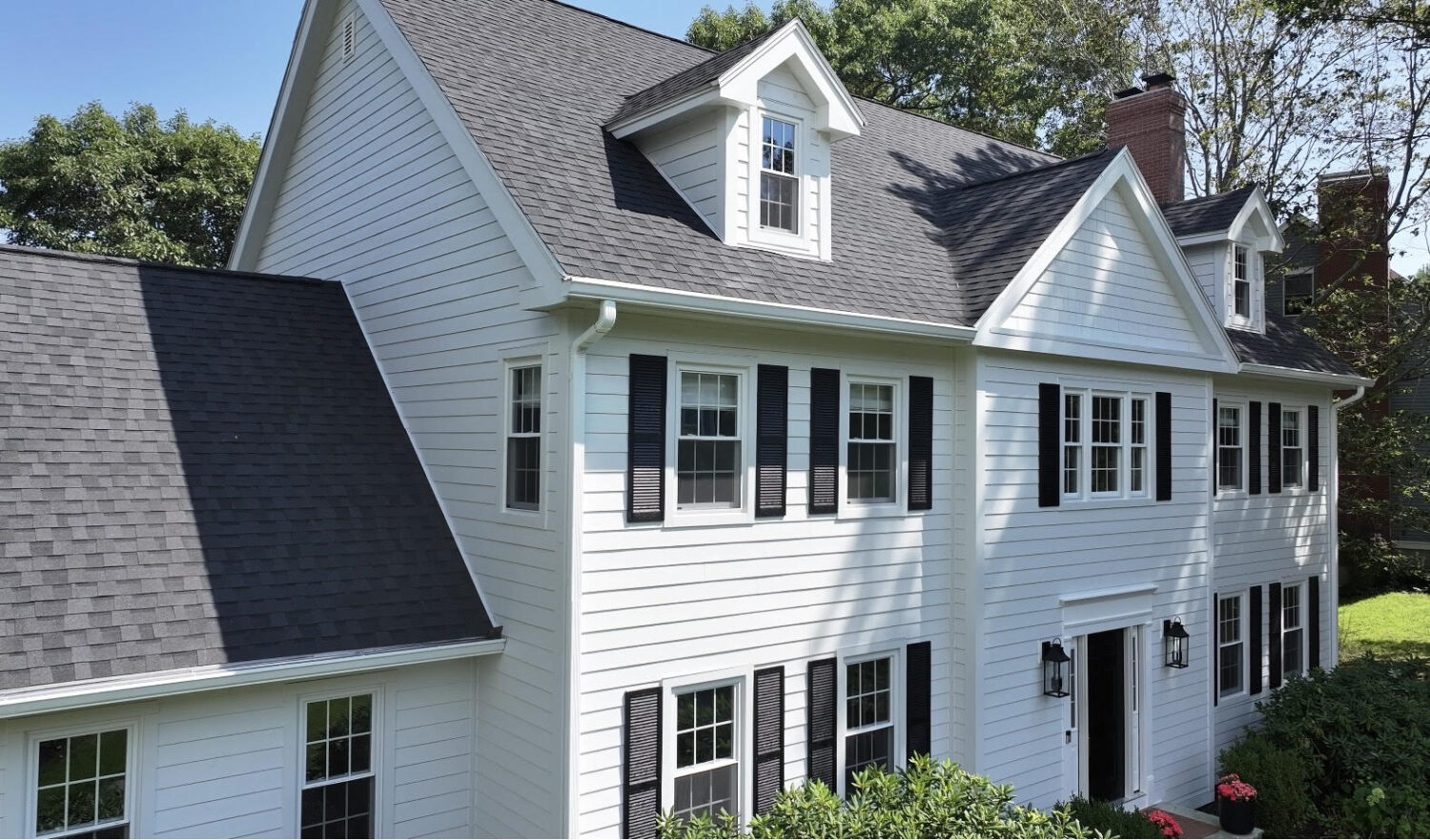 Large white colonial-style house with black shutters and a dark grey roof. It has a symmetrical façade with multiple windows and a prominent central entrance. Surrounded by greenery and trees under a clear blue sky.