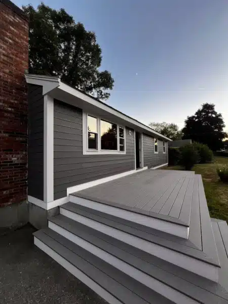 A modern house in Newburyport, MA, showcases a stylish James Hardie Installation with dark gray siding. It features a newly built, spacious light gray deck with clean lines and white trim. A brick chimney is visible on the left against the dusk sky, framed by trees in the background.