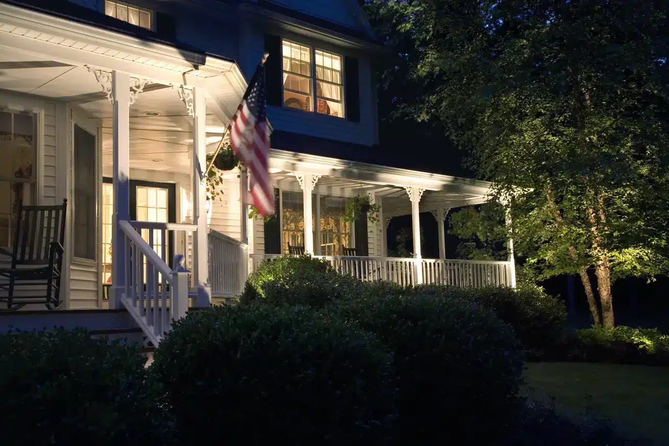 A warmly lit, two-story house at night features exquisite outdoor lighting design with a wraparound porch. An American flag hangs by the steps, while bushes fringe the porch and trees stand proudly to the right. The windows glow invitingly, casting a cozy atmosphere all around.