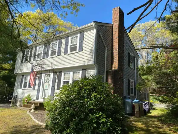 A two-story house with light gray vinyl siding and white trim rests in a lush green yard. The red brick chimney adds charm, while an American flag waves by the front door. Under a clear blue sky, its surrounded by tall trees, inviting thoughts of Can I paint vinyl siding?.