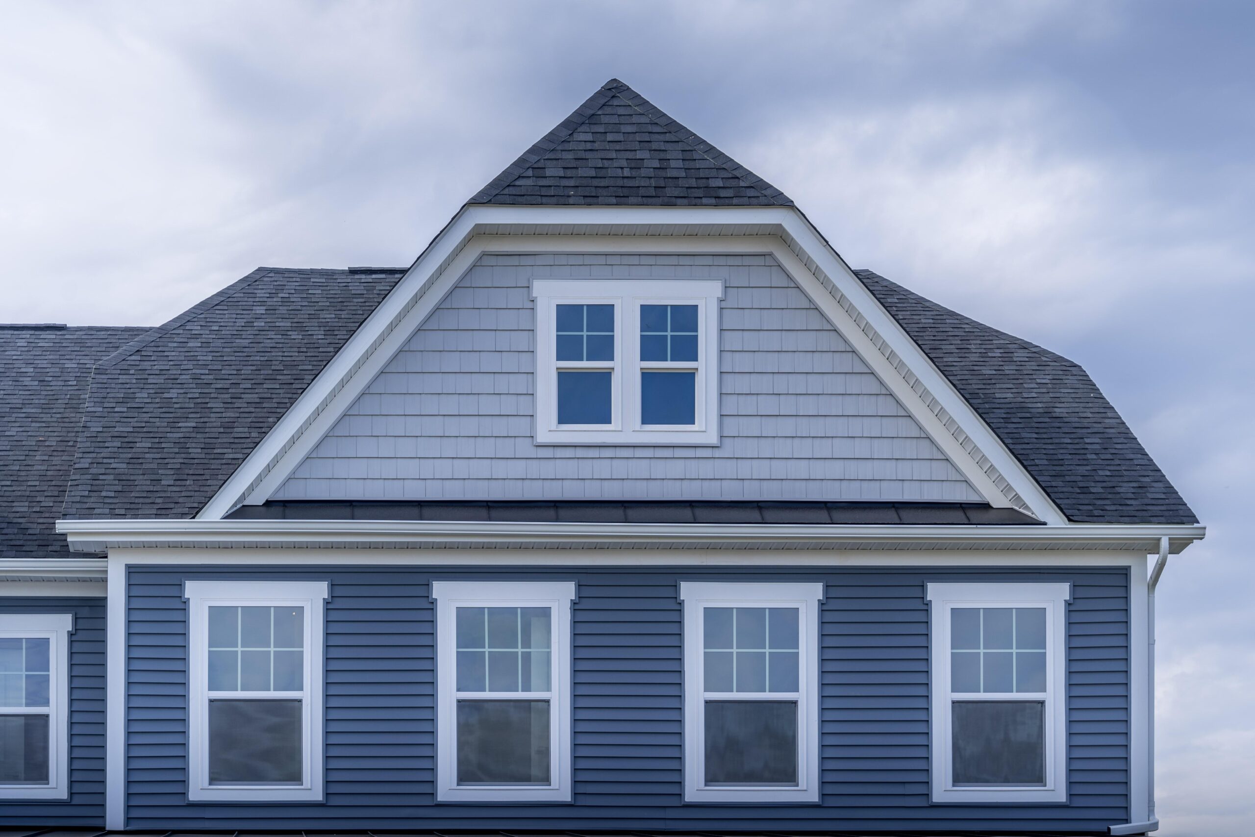 The upper portion of a modern house with gray siding, white trim, symmetrical Replacement Windows in Newbury MA, and a steeply pitched roof under a cloudy sky.