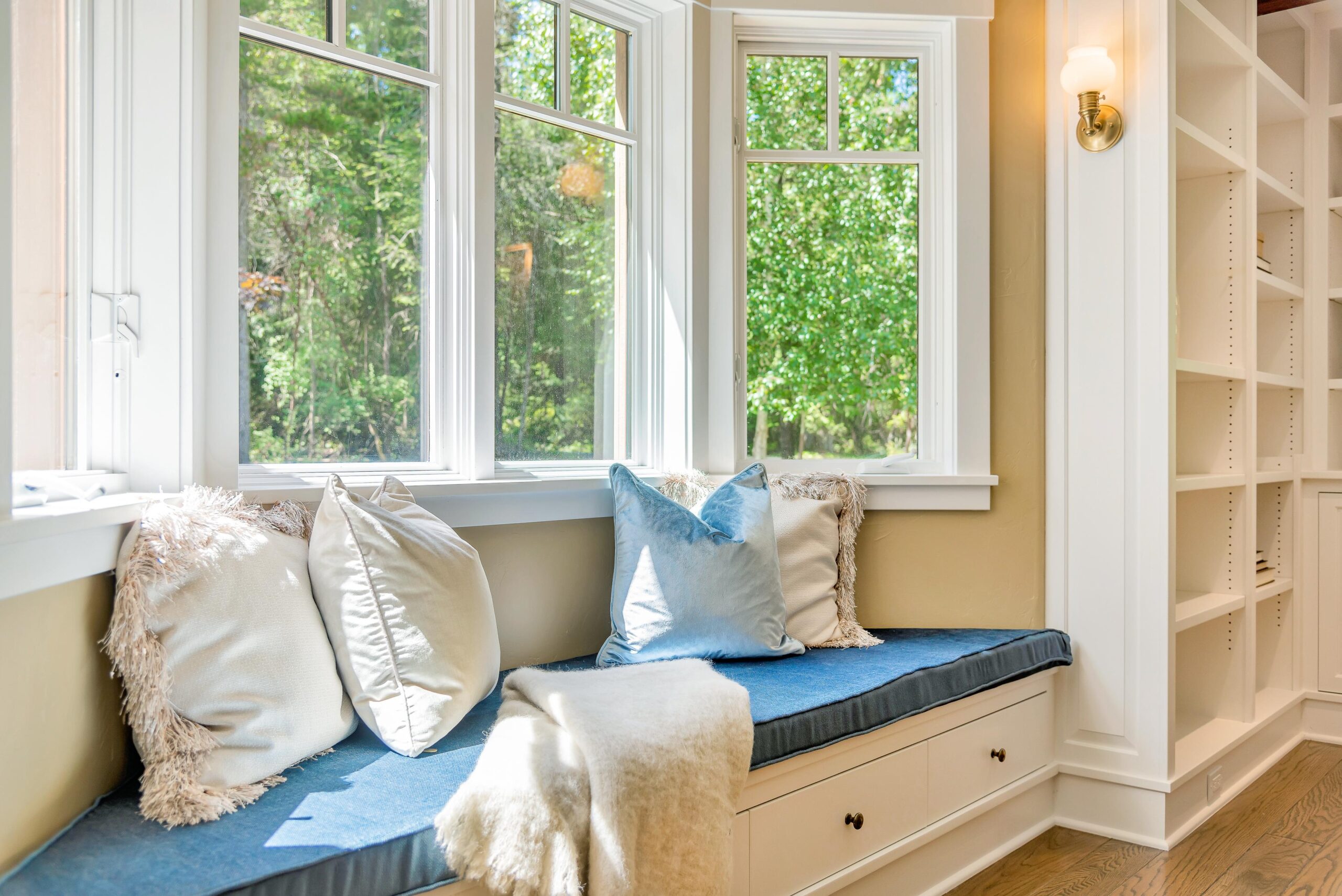 A cozy window seat with blue cushions, white and light blue pillows, and a cream throw blanket sits beside large replacement windows in Newbury MA, overlooking green trees. Built-in shelves and a wall sconce are on the right. Sunlight fills the space.