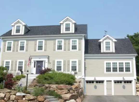 Two-story beige house with white trim, three dormer windows, and attached garage showcases expert roofing in Acton MA, plus a landscaped front yard with rocks, shrubs, and an American flag by the front porch.