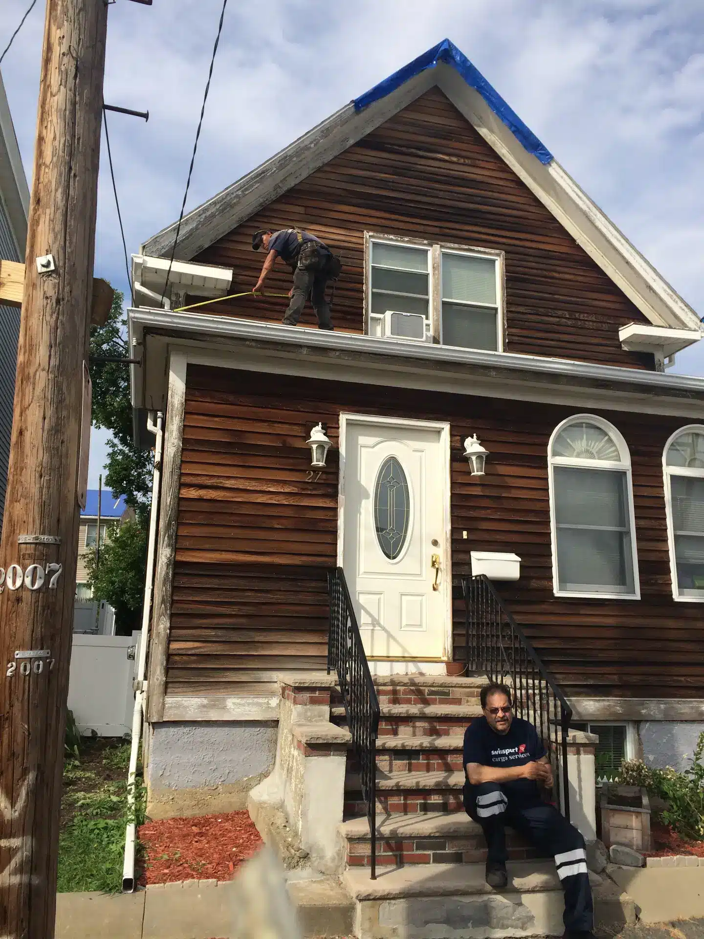 A person stands on the roof edge working near a window of a wooden house—this week’s Wednesday Weekly Wow Roof Rescue Winner—while another person sits on the front steps below, looking at a phone. The house has brown siding and white trim.