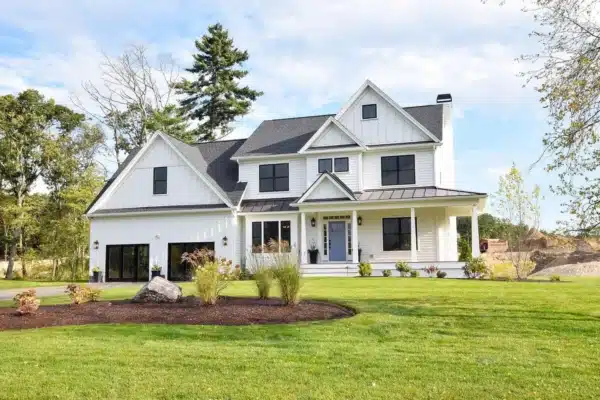 A two-story white modern farmhouse with the best siding for a house boasts a dark gray roof amidst a green lawn. It features black window frames, a front porch, landscaped shrubs, and trees in the background under a clear blue sky.