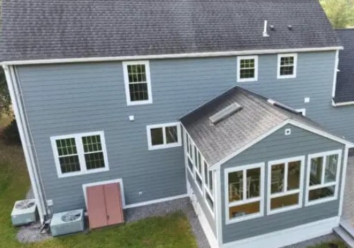 A gray house with white trim features a sunroom extension with replacement windows in Methuen MA, a skylight on the roof, and two air conditioning units beside pink doors outside on a grassy yard.