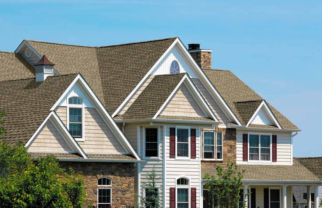 A large house with multiple gabled roofs, featuring beige siding and red shutters. The walls blend stone and siding, while brown shingles cover the roof equipped with a modern roofing ventilation system. Greenery and a blue sky complete the scene.