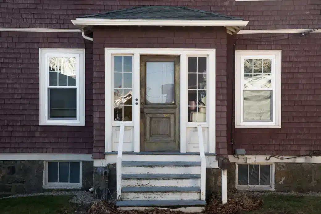 A small wooden porch with white railings and steps leads to a weathered front door, where leaking air may slip through, flanked by two tall windows on a maroon-sided house with stone foundation.