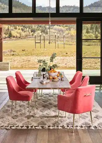 A dining room with a rectangular table, six pink upholstered chairs, and a light-patterned rug. Large windows reveal scenic green fields and hills. A bowl of fruit on the table adds a touch of Fall Inspiration to the home decor.