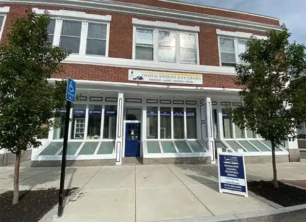 A two-story brick building with large windows displays signs for Coastal Windows & Exteriors, specializing in home exterior maintenance. The storefront features a blue door, and there is a handicap parking sign in front. Trees are planted along the sidewalk.