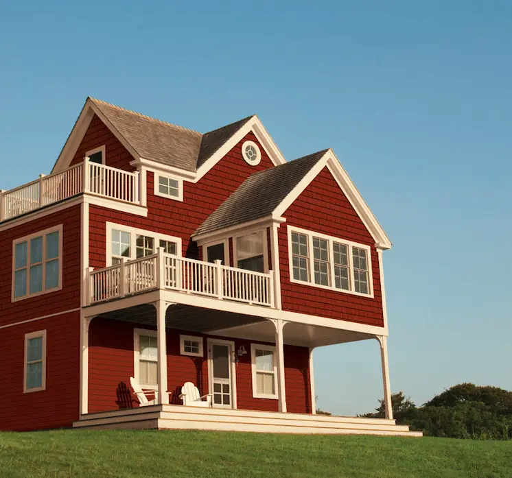A charming two-story red house with durable siding and white trim features a porch and a balcony. It boasts large windows beneath its pitched roof. Two white chairs rest on the porch. The home is beautifully set on a grassy hill under a clear blue sky.