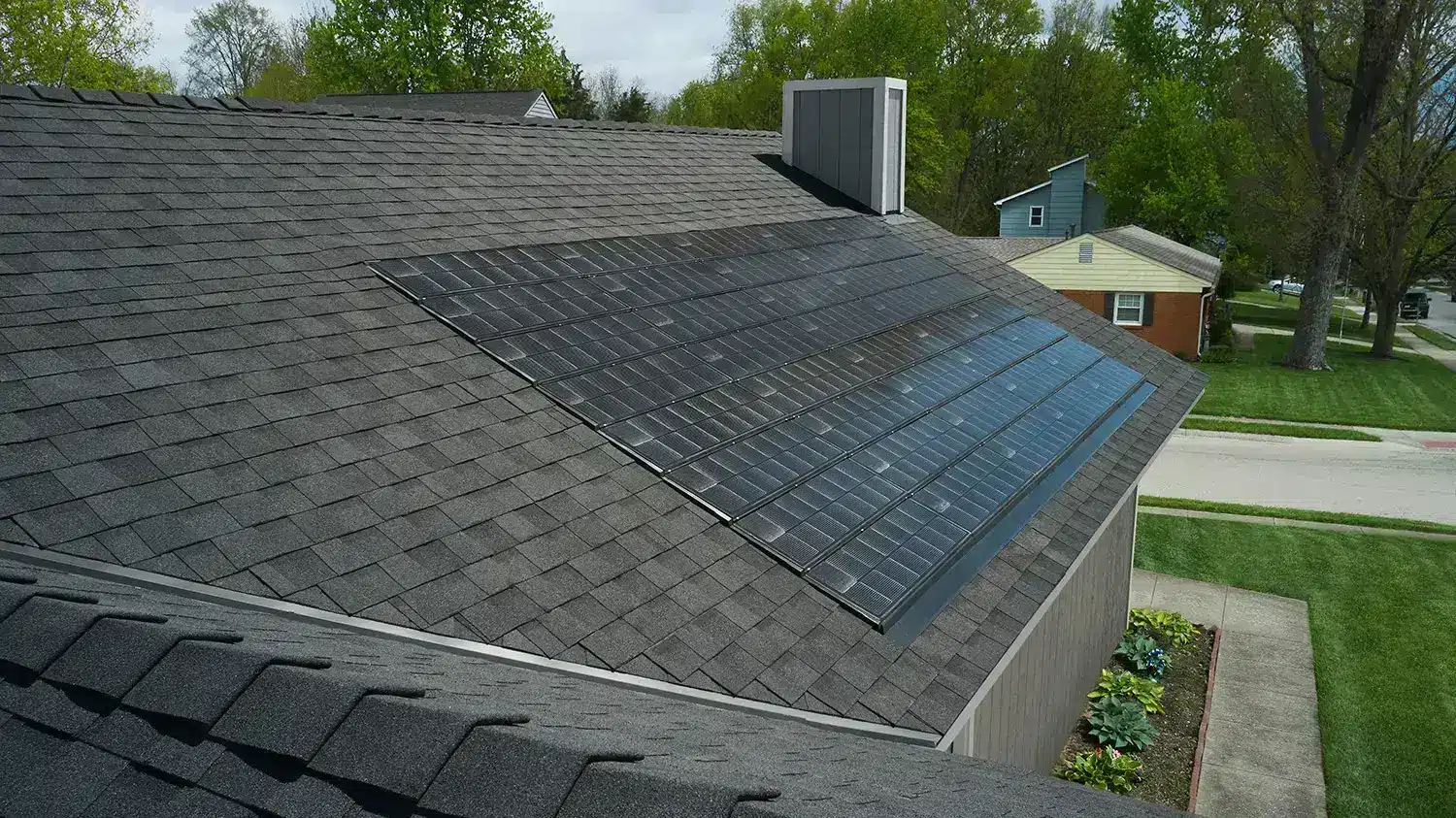 A house with dark shingles and solar panels on its roof showcases an eco-friendly touch, raising curiosity about what solar shingles are. The tidy neighborhood features well-maintained lawns and trees, while another house stands in the background with a paved road and a sidewalk alongside.