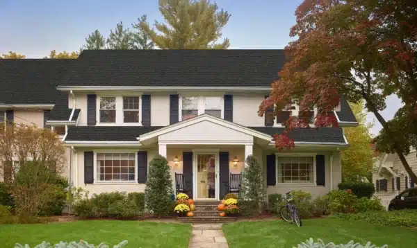 A two-story house with a black roof and white exterior features blue shutters and a small porch adorned with pumpkins for autumn. As cold weather approaches, ensure fall window maintenance is completed. Two chairs and a bicycle sit by the entrance, surrounded by red-leaved trees on the green lawn.