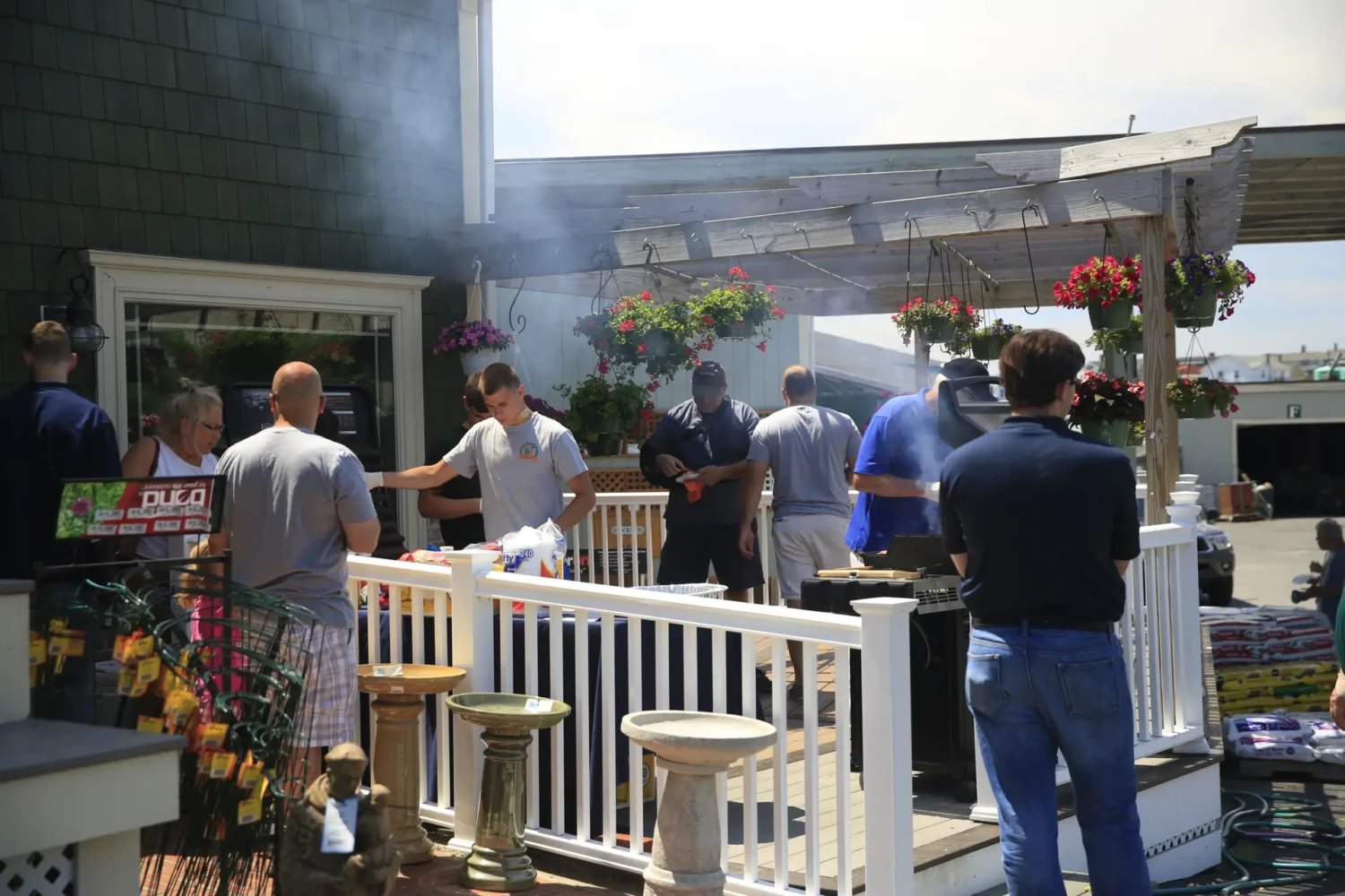 A group of people gather on a sunny deck for a barbecue, surrounded by hanging flower baskets and outdoor decor, while smoke rises from the grill—true Passport to Summer Living event photos.