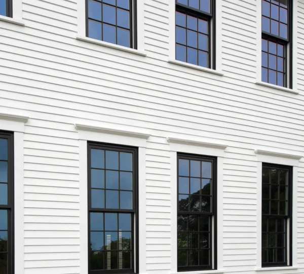 A white building with horizontal siding features three sets of double-hung windows, each with multiple panes. The black frames contrast with the white exterior, expertly designed to stop windows from leaking water. Reflections of trees and sky can be seen in the window glass.