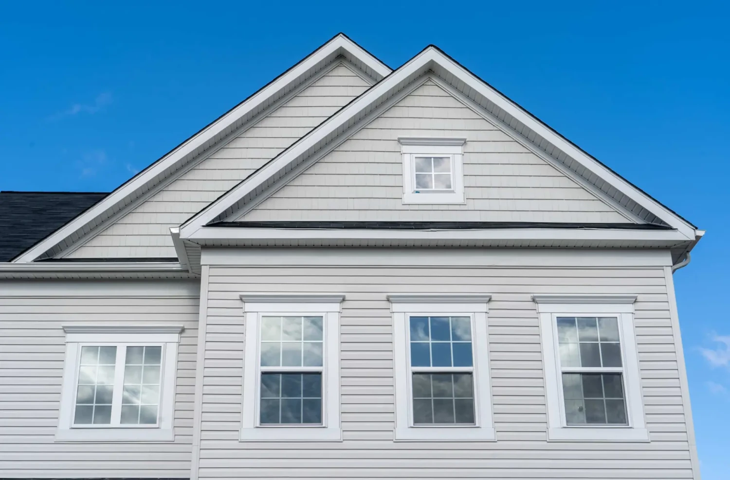 A two-story house with gray siding and a black shingle roof stands against a clear blue sky. Featuring symmetrical rectangular windows and a triangular gable above, its an inviting sight. Considering new windows could further enhance the homes value and appeal.