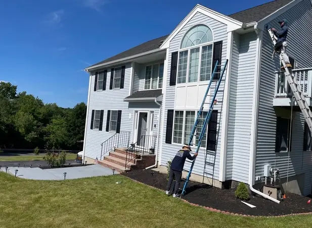 Two individuals on ladders diligently follow a window replacement checklist as they work on the exterior of a two-story house with light gray siding and black shutters. The house features a large arched window and a cozy front porch with steps. Above, a clear sky stretches over the grassy surroundings.