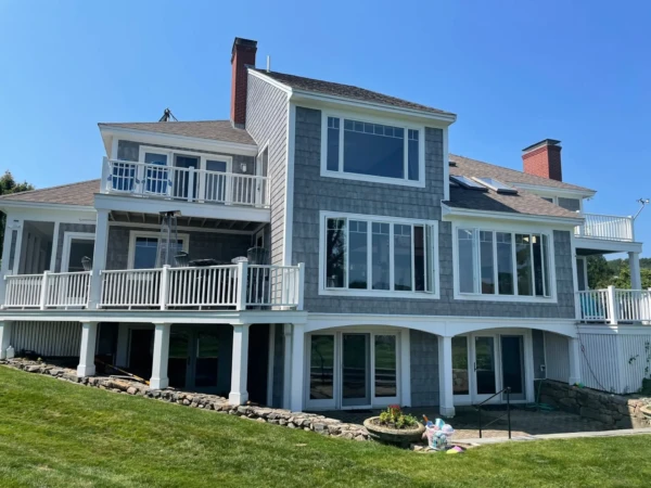 A large, two-story gray house with white trim boasts undeniable curb appeal. Featuring a spacious balcony, numerous large windows, and two chimneys, its surrounded by lush green grass and a stone retaining wall. A white fence encloses the scenic balcony area under a clear blue sky.