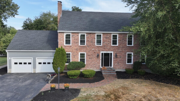 A charming two-story brick house in North Andover, MA, boasting a dark shingle roof. It features six windows on the second floor and four on the first, with a black double front door. The house includes an adjacent garage with James Hardie Installation and a landscaped yard where a bicycle stands nearby.