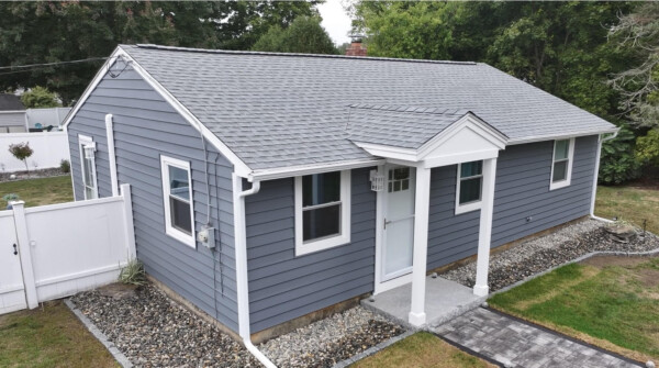 A charming small gray house in Hampton, NH, features a gabled roof and white trim. Surrounded by a neatly landscaped yard with stones bordering the home, the property includes vinyl siding installation for durability. A white fence is visible with trees elegantly enclosing the scene.