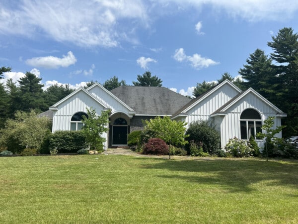 A single-story house in Plymouth, MA, boasts pristine white vinyl siding and a dark roof, surrounded by lush bushes and trees. The front yard features a well-maintained green lawn under a blue sky with scattered clouds.