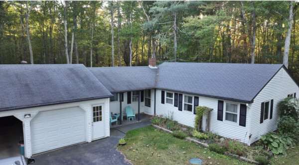 Aerial view of a charming single-story white house with vinyl siding installation and a dark gray roof, nestled amidst the tall trees in Wells, ME. It features a garage on the left and a small garden out front. Two blue chairs adorn the porch near the entrance.