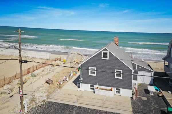 A gray wooden house with a chimney exudes coastal curb appeal near a sandy beach with gentle waves. Decorative items line the pathway to the sea, and a paved driveway is in the foreground. A sunny, clear blue sky completes this charming seaside scene.