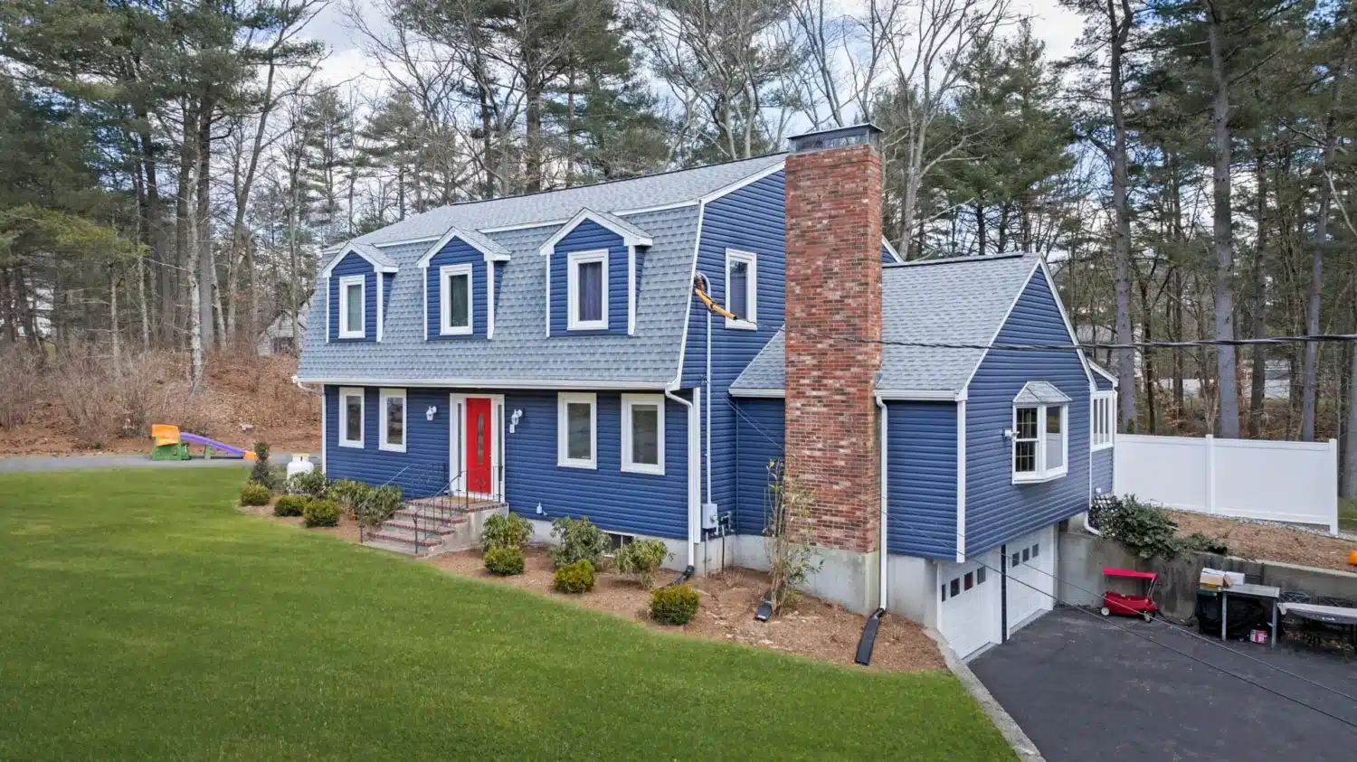 A two-story blue house with a gray roof in good condition and a red front door features multiple windows and a prominent brick chimney. The house is surrounded by a manicured lawn, bordered by trees, with a driveway leading to the garage, showing no current need to repair vs replace the roof.