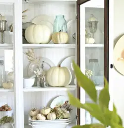 White cabinet with glass door showcasing autumn decor: white pumpkins, a blue mason jar, plates, and dried plants. A green plant is partially visible in the foreground. The overall theme is rustic and cozy, inviting you to embrace Sweet Autumn at home.