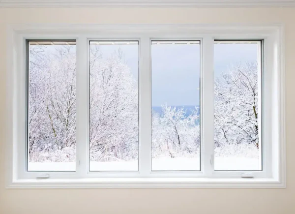 Through the energy-efficient windows in MA, a four-pane view reveals a snowy landscape with bare, snow-covered trees against a pale, cloudy sky. The rooms simple interior features light-colored walls and a pristine white window frame.