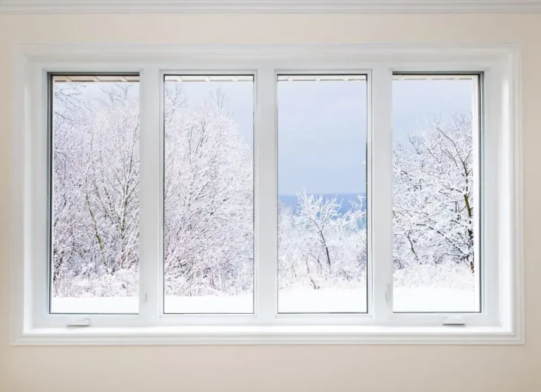 Through the energy-efficient windows in MA, a four-pane view reveals a snowy landscape with bare, snow-covered trees against a pale, cloudy sky. The rooms simple interior features light-colored walls and a pristine white window frame.