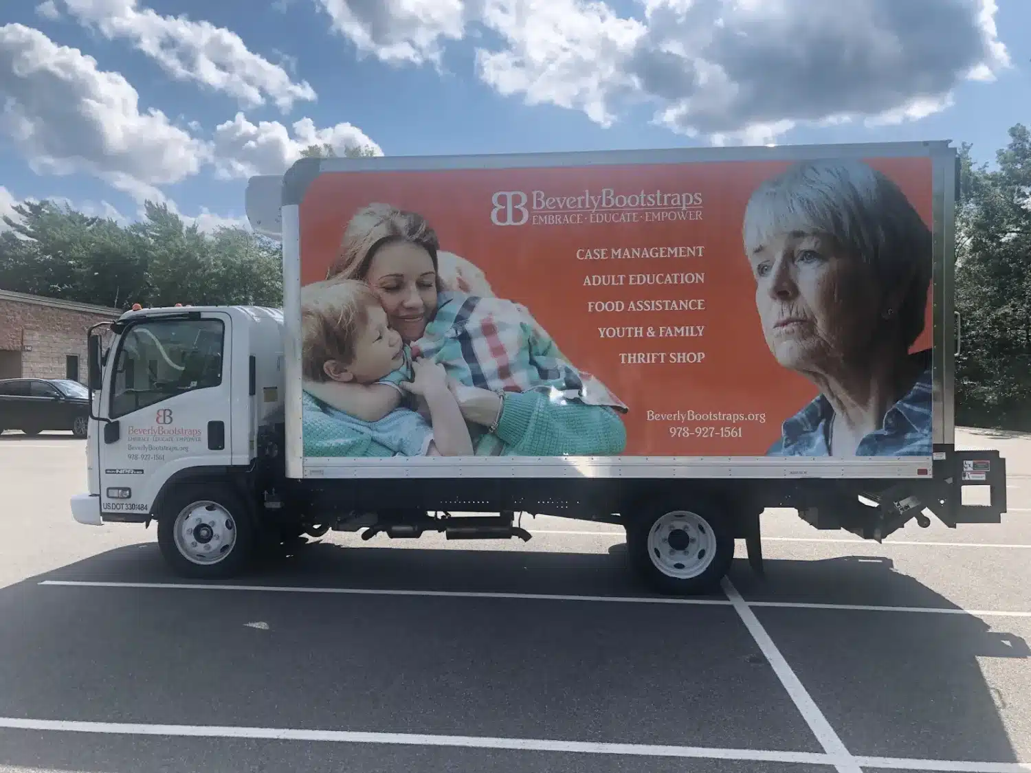 A truck parked in a lot displays an advertisement for Beverly Bootstraps in Massachusetts. The ad shows a cheerful woman with a child, and an elderly woman looking ahead. Services listed include case management, food assistance, and a thrift shop—supporting the community through COVID-19 challenges.