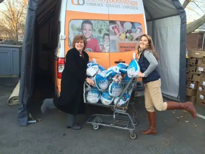 Two women smile while posing with a shopping cart full of frozen turkeys for a Thanksgiving charity event, in front of a Beverly Patch donation van. Both have one foot playfully lifted and appear happy and cheerful.