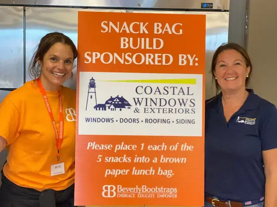 Two smiling women stand beside a sign reading, Snack Bag Build Sponsored By: Coastal Windows & Exteriors. Help us deliver 750 healthy snacks to children—please place one of each snack into a brown paper lunch bag.
