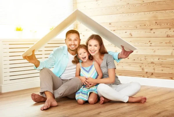 A happy family of three sits on a wooden floor under a white cardboard roof. The parents, smiling as they hold the roof over their heads, seem to say their current roofing needs no replacement for now. Their young daughter sits between them, grinning in the cozy interior setting.