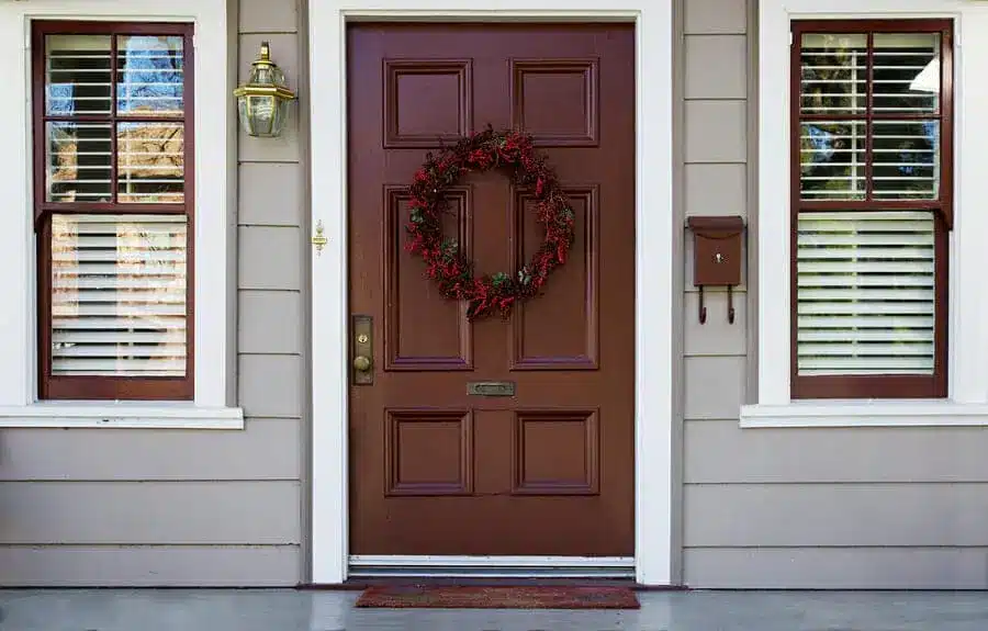 A brown entry door with a red berry wreath is centered between two windows with white shutters. A brass light fixture graces the left, while a small brown mailbox is mounted on the right side, creating a charming entrance.