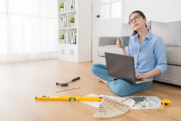 A woman sits cross-legged on the floor with a laptop, holding a mug and looking thoughtful. Surrounding her are a level tool, paint swatches, and papers hinting at a cost vs. value deliberation. A sofa and bookshelves add to the ambiance of her home renovation project.