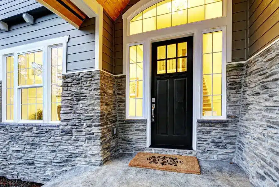 Front entryway of a house with striking entry doors, large windows on either side, gray stone exterior, and a “Welcome” doormat on a concrete porch. Warm light glows from inside the house.