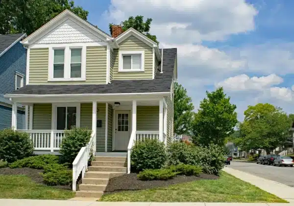 A two-story house with light green vinyl siding, white trim, and a covered porch showcases its charm. It features a gabled roof and a small flight of steps leading to the front door. The yard is landscaped with shrubs, and there are trees and neighboring houses in the background.