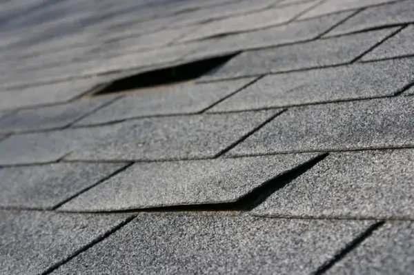 Close-up of a gray asphalt shingled roof with several shingles lifted or missing, revealing gaps. The surface appears weathered, highlighting spring home maintenance needs. The angle emphasizes the unevenness and urgency for repair.