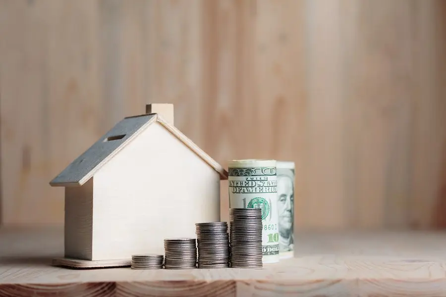 A small wooden house model sits next to stacks of coins and a roll of U.S. dollar bills on a wooden surface, symbolizing real estate and home improvement financing. The background, a blurred wood panel texture, subtly enhances the theme of financial investment.